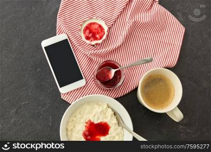 food and breakfast concept - porridge in bowl, smartphone, jam in mason jar, spoon and cup of coffee on slate stone table. porridge with jam, spoon, coffee and phone