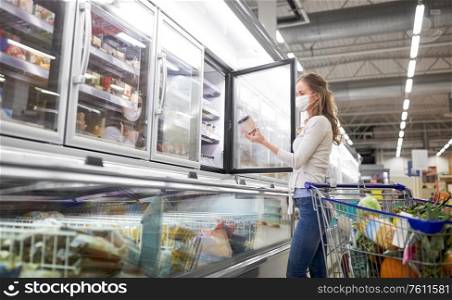 food and pandemic concept - woman in face protective medical mask for protection from virus disease with shopping cart choosing ice cream at grocery store freezer. woman in mask choosing ice cream at grocery store