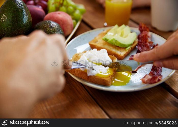 food and people concept - hands of man eating toast with pouched egg, avocado and bacon with fork and knife. man eating toast with pouched egg and bacon
