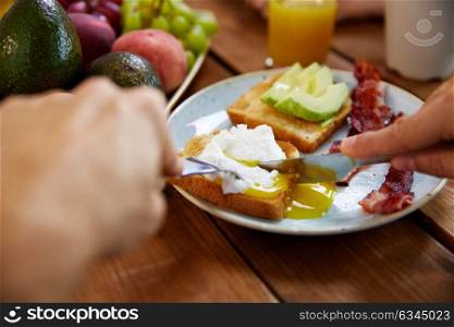 food and people concept - hands of man eating toast with pouched egg, avocado and bacon with fork and knife. man eating toast with pouched egg and bacon