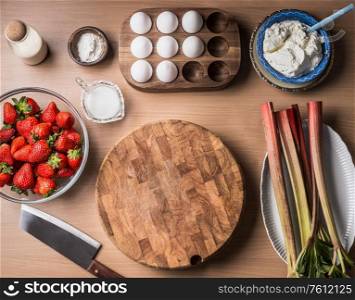 Food background frame with strawberries and rhubarb , eggs, cottage cheese around wooden cutting board. Top view. Seasonal cooking and baking. Healthy clean organic food.