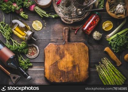 Food background. Various ingredients and seasonings for tasty cooking around cutting board on wooden rustic table. Oil,lemon, vine bottle, pickled chili in jar, asparagus, herbs and spices. Frame
