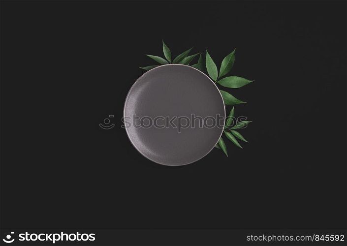 Food background with a grey empty plate with green leaves ornament on a black table. Above view of blank tableware. Flat lay with an empty dish.