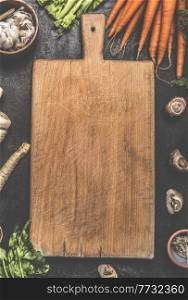 Food background with blank wooden cutting board, carrots, mushrooms and garlic on dark kitchen table. Cooking preparation at home. Top view with copy space.