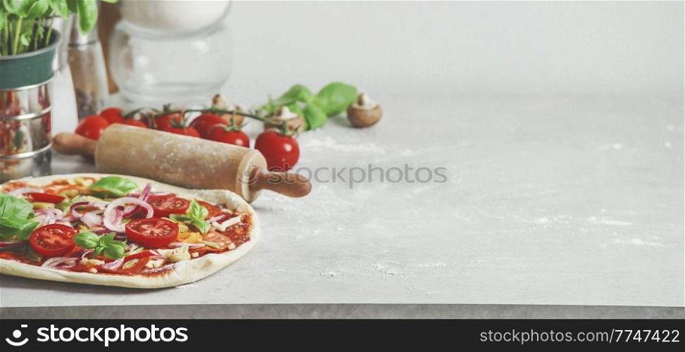 Food background with homemade Italian pizza, basil, tomatoes and wooden rolling pin on pale grey kitchen table. Cooking at home with fresh ingredients. Front view with copy space.