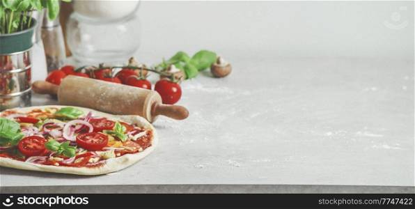 Food background with homemade Italian pizza, basil, tomatoes and wooden rolling pin on pale grey kitchen table. Cooking at home with fresh ingredients. Front view with copy space.