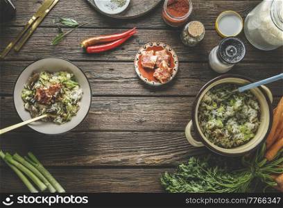 Food background with rice dish in bowl with leek and meat, vegetables, chili pepper, herbs, spices, cutlery and kitchen utensils at rustic brown wooden table. Cooking healthy food. Top view.
