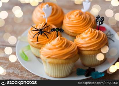 food, baking and holidays concept - cupcakes or muffins with halloween party decorations and candies on plate. halloween party decorated cupcakes on plate