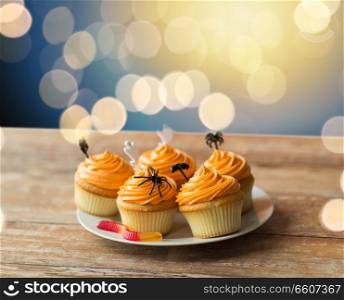 food, baking and holidays concept - cupcakes or muffins with halloween party decorations and candies on plate over lights. halloween party decorated cupcakes on plate