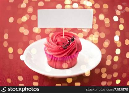 food, baking and pastry concept - close up of cupcake or muffin on plate with red buttercream frosting and blank nametag over festive lights. close up of cupcake with red buttercream frosting