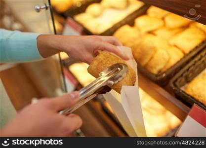 food, baking and sale concept - close up of hand with tongs and paper bag buying bun at bakery or grocery store