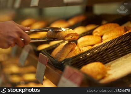 food, baking and sale concept - close up of hand with tongs taking bun at bakery or grocery store