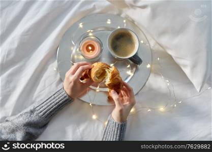 food, breakfast and people concept - hands of woman eating croissant with coffee in bed. hands of woman eating croissant with coffee in bed