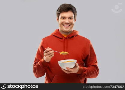 food, breakfast and people concept - smiling young man in red hoodie eating cereals over grey background. smiling young man in red hoodie eating cereals