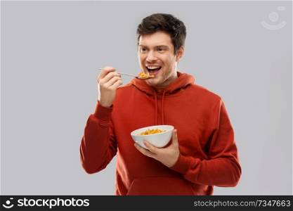 food, breakfast and people concept - smiling young man in red hoodie eating cereals over grey background. smiling young man in red hoodie eating cereals