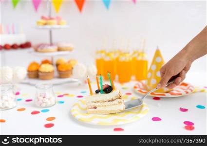 food, celebration and festive concept - hand with shovel putting piece of cake with candles on plate at birthday party. hand putting piece of birthday cake on plate