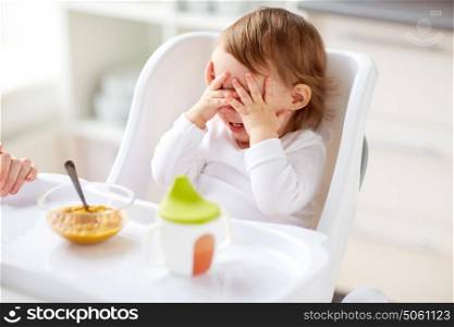 food, child, feeding and people concept - baby with spoon sitting in highchair and eating puree from at home kitchen. baby sitting in highchair and eating at home