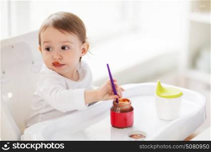 food, child, feeding and people concept - baby with spoon sitting in highchair and eating puree from jar at home kitchen. baby with spoon eating puree from jar at home