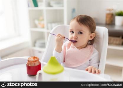 food, child, feeding and people concept - little baby girl with spoon sitting in highchair and eating puree from jar at home kitchen. baby girl with spoon eating puree from jar at home