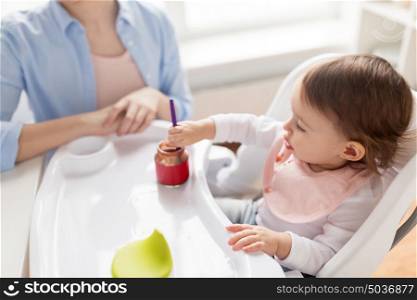 food, child, feeding and people concept - little baby girl with spoon sitting in highchair and eating puree from jar and mother at home kitchen. baby girl with spoon eating puree from jar at home