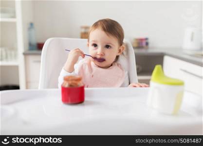 food, child, feeding and people concept - little baby girl with spoon sitting in highchair and eating puree from jar at home kitchen. baby girl with spoon eating puree from jar at home