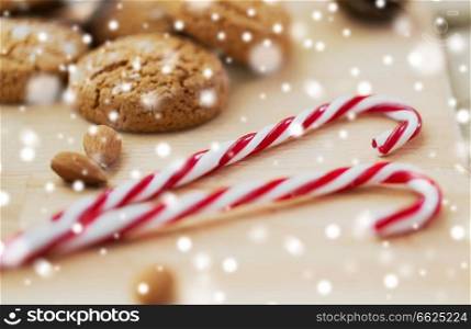 food, christmas and eating concept - close up of candy canes and oatmeal cookies with almonds on wooden board over snow. candy canes, cookies and almonds on board