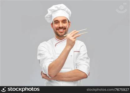 food cooking, asian cuisine and people concept - happy smiling male chef in toque holding something with chopsticks over grey background. happy smiling male chef with chopsticks