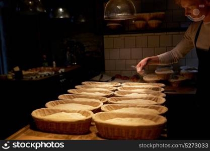 food cooking, baking and people concept - baker making bread and pouring flour to baskets while dough rising at bakery kitchen. baker with baskets for dough rising at bakery