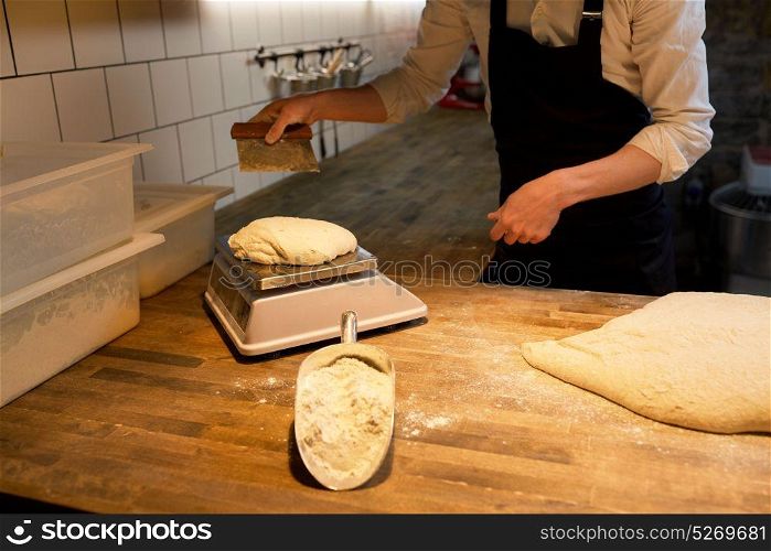 food cooking, baking and people concept - baker with bench cutter portioning and weighing bread dough on scale at bakery kitchen. baker weighing bread dough on scale at bakery