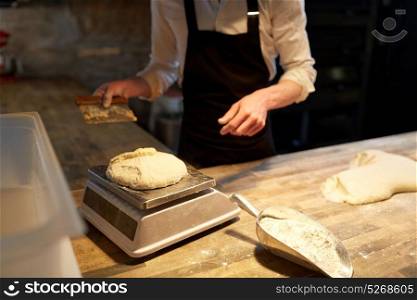 food cooking, baking and people concept - chef or baker with bench cutter portioning and weighing dough on scale at bakery. chef or baker weighing dough on scale at bakery
