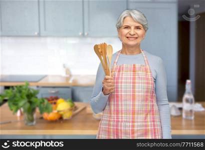 food cooking, culinary and old people concept - portrait of smiling senior woman in apron with wooden spoons over kitchen background. smiling senior woman in apron with wooden spoons