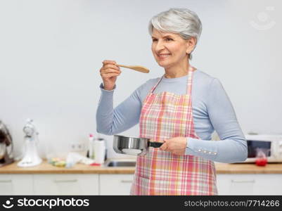 food cooking, culinary and old people concept - portrait of smiling senior woman in apron with pot and spoon over kitchen background. senior woman in apron with pot cooking food