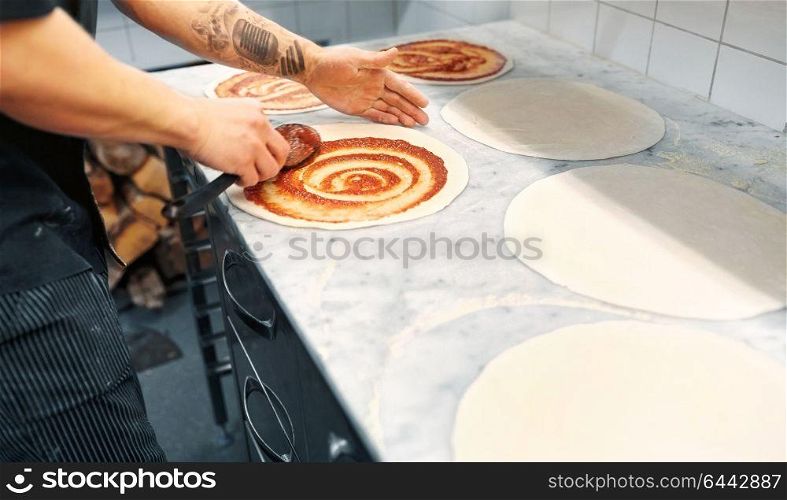 food cooking, culinary and people concept - cook or baker with spoon applying tomato sauce to raw pizza dough at pizzeria. cook applying tomato sauce to pizza at pizzeria