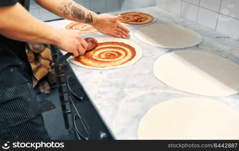 food cooking, culinary and people concept - cook or baker with spoon applying tomato sauce to raw pizza dough at pizzeria. cook applying tomato sauce to pizza at pizzeria