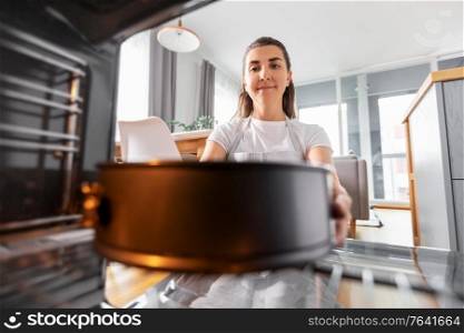 food cooking, culinary and people concept - young woman putting baking dish into oven at home kitchen. woman cooking food in oven at home kitchen