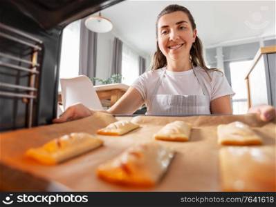 food cooking, culinary and people concept - young woman putting baking tray with pies into oven at home kitchen. woman cooking food in oven at home kitchen