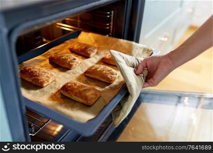 food cooking, culinary and people concept - young woman with potholder taking baking tray with jam pies out of oven at home kitchen. woman cooking food in oven at home kitchen