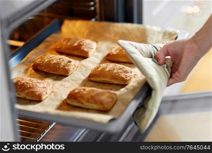 food cooking, culinary and people concept - young woman with potholder taking baking tray with jam pies out of oven at home kitchen. woman cooking food in oven at home kitchen