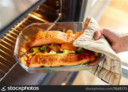 food cooking, culinary and people concept - young woman with potholder taking baking dish with salmon fish and vegetables out of oven at home kitchen. woman cooking food in oven at home kitchen