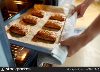 food cooking, culinary and people concept - young woman with towel taking baking tray with jam pies out of oven at home kitchen. woman cooking food in oven at home kitchen