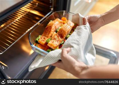 food cooking, culinary and people concept - young woman with towel taking baking dish with salmon fish and vegetables out of oven at home kitchen. woman cooking food in oven at home kitchen