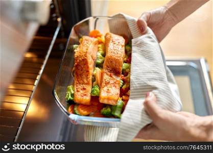 food cooking, culinary and people concept - young woman with towel taking baking dish with salmon fish and vegetables out of oven at home kitchen. woman cooking food in oven at home kitchen
