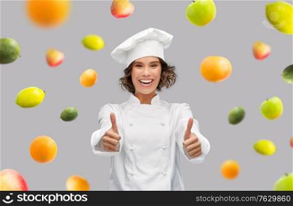 food cooking, gesture and people concept - happy smiling female chef in toque showing thumbs up over fruits on grey background. happy female chef showing thumbs up over fruits