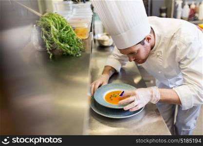 food cooking, profession and people concept - happy male chef cook serving and decorating plate of soup with pansy flower at restaurant kitchen. male chef decorating dish with pansy flower