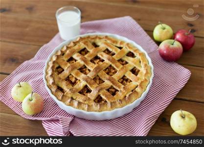 food, culinary and baking concept - apple pie, glass of milk and kitchen towel on wooden table. apple pie in baking mold on wooden table