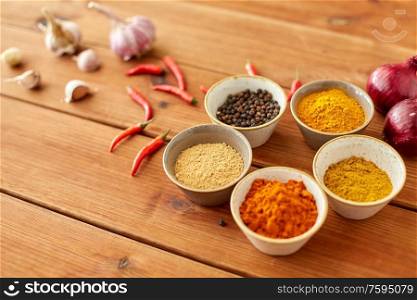 food, culinary and eating concept - bowls with different spices, onion, garlic and red hot chili peppers on wooden table. spices, onion, garlic and red hot chili peppers
