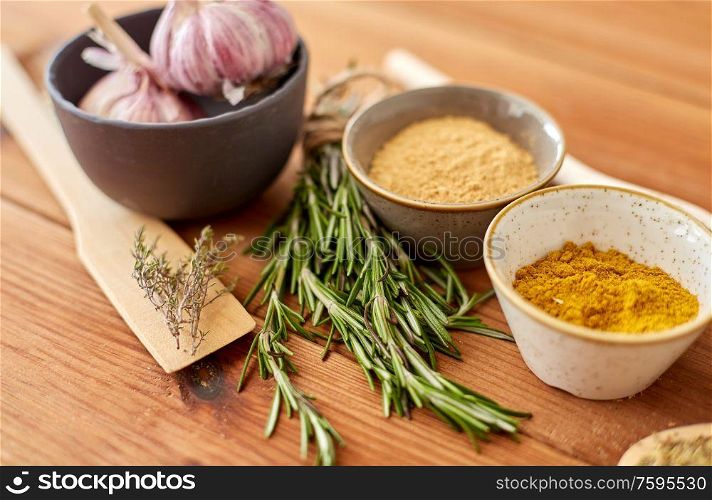 food, culinary and eating concept - bowls with different spices, rosemary, spatula and garlic on wooden table. spices, rosemary, wooden spatula and garlic