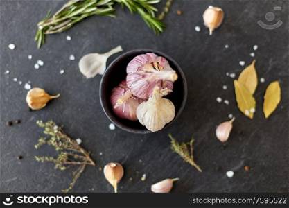 food, culinary and eating concept - garlic in bowl, rosemary and salt on stone surface. garlic in bowl and rosemary on stone surface