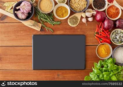 food, culinary and eating concept - tablet pc computer among different spices, onion, garlic with pine nuts and red hot chili peppers on wooden table. tablet pc computer among spices on wooden table
