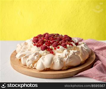 food, culinary, baking and cooking concept - close up of pavlova meringue cake decorated with red berries on wooden serving board and kitchen towel over yellow wall background. pavlova meringue cake with berries on wooden board
